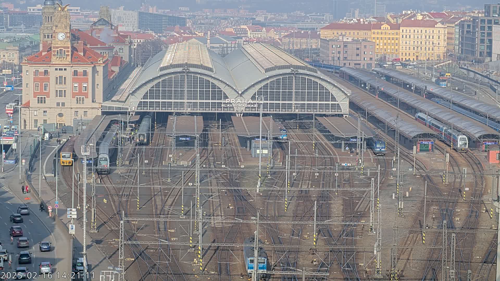 A large train station with numerous tracks and trains, situated in a city under a clear, sunny sky.