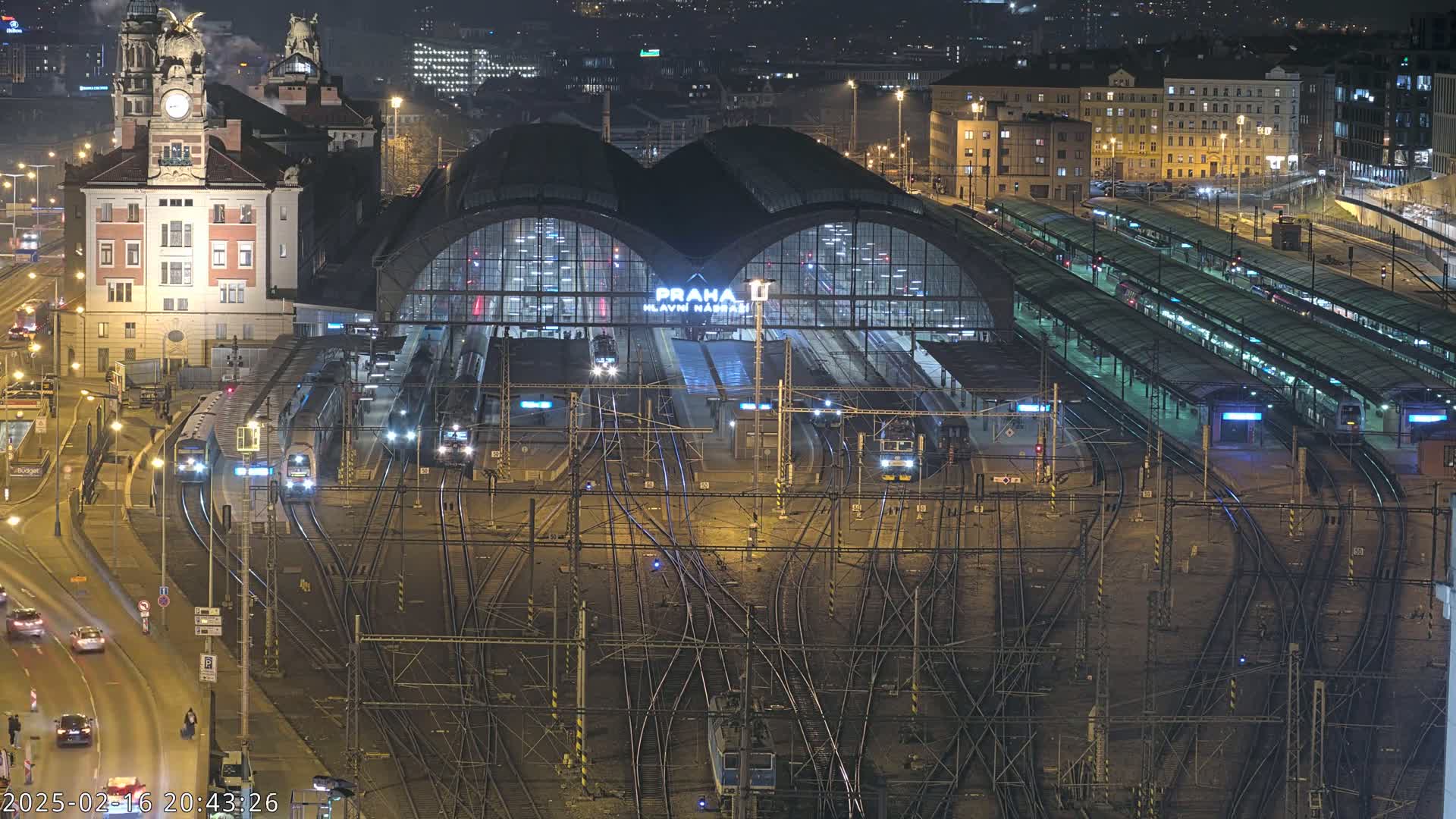 A large, illuminated train station at night with numerous trains and tracks, surrounded by city buildings.