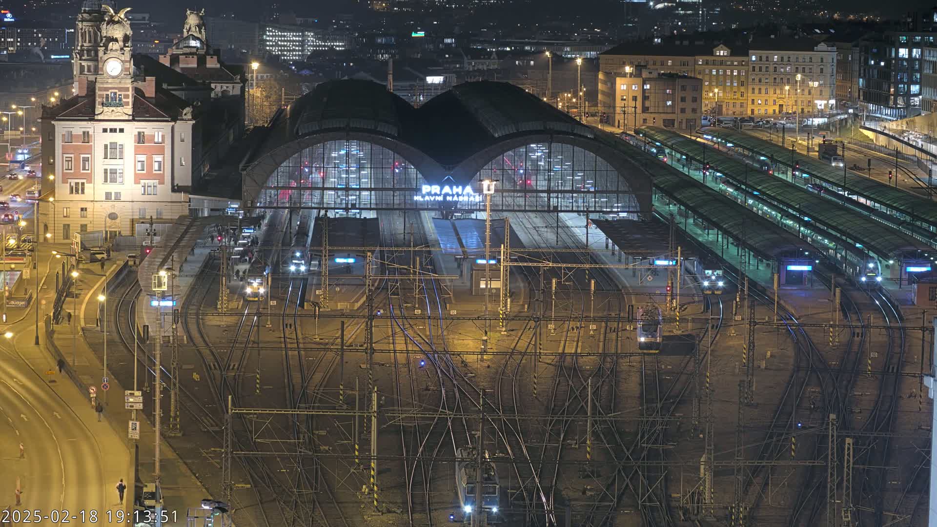 A nighttime aerial view of a large, illuminated train station with many tracks and platforms, surrounded by city lights.