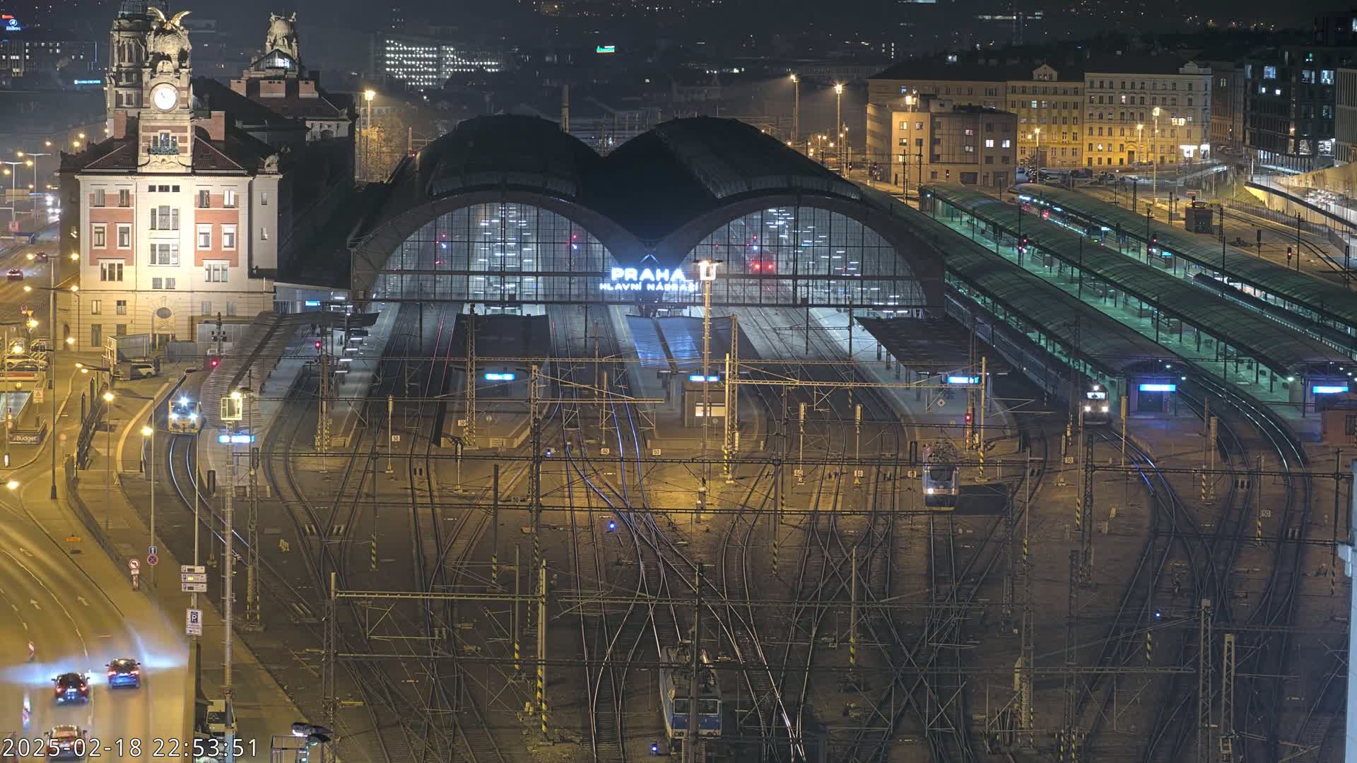 A nighttime aerial view of a large train station with numerous tracks and platforms, surrounded by city buildings and illuminated streets.