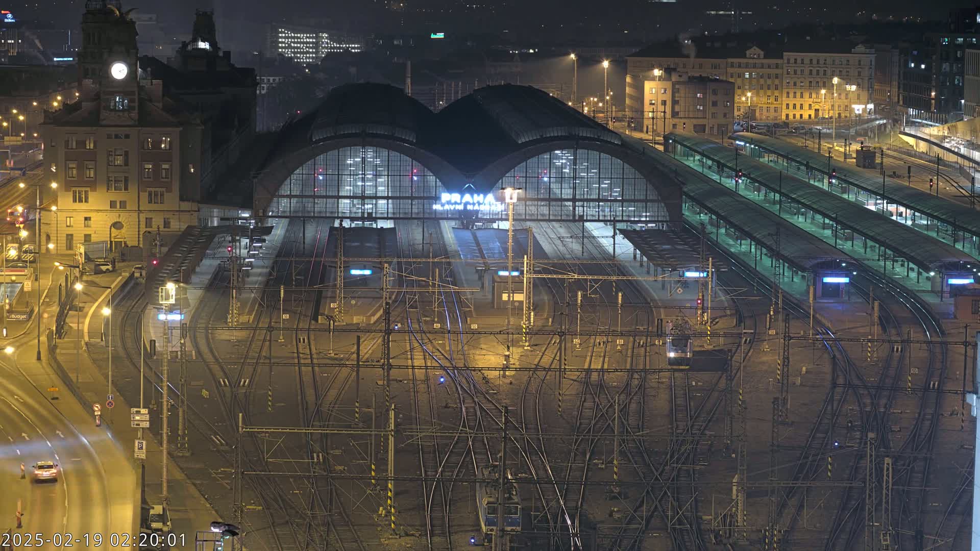 A large, illuminated train station at night, surrounded by a complex network of train tracks and platforms, with city buildings visible in the background under a dark sky.