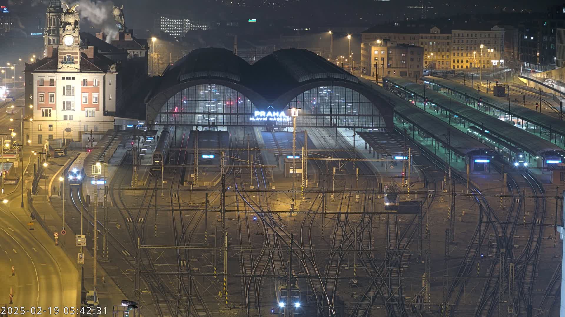 A nighttime aerial view of a large, illuminated train station with numerous tracks and trains, under clear skies.