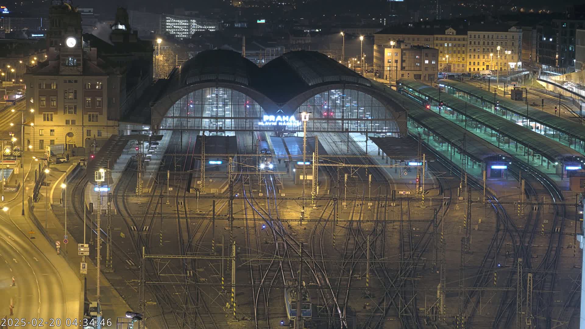 A nighttime aerial view of a large, illuminated train station with numerous tracks and platforms, surrounded by city buildings.