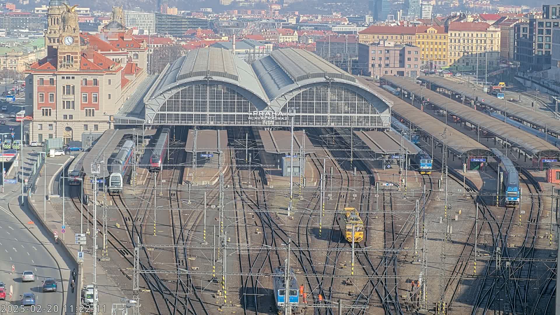 A large train station with numerous tracks and trains, under a sunny sky, is surrounded by city buildings.