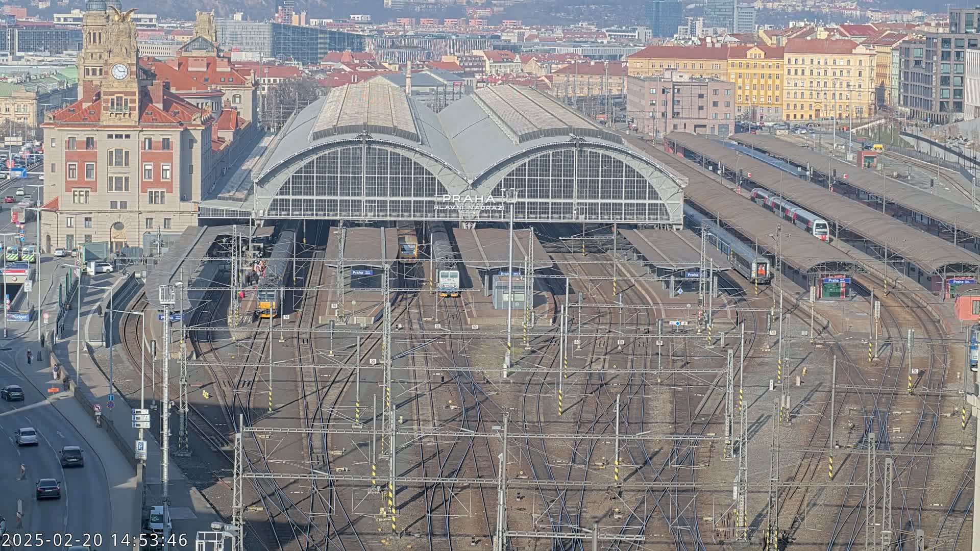 A large train station with numerous tracks and trains, under a clear sunny sky, is viewed from above.