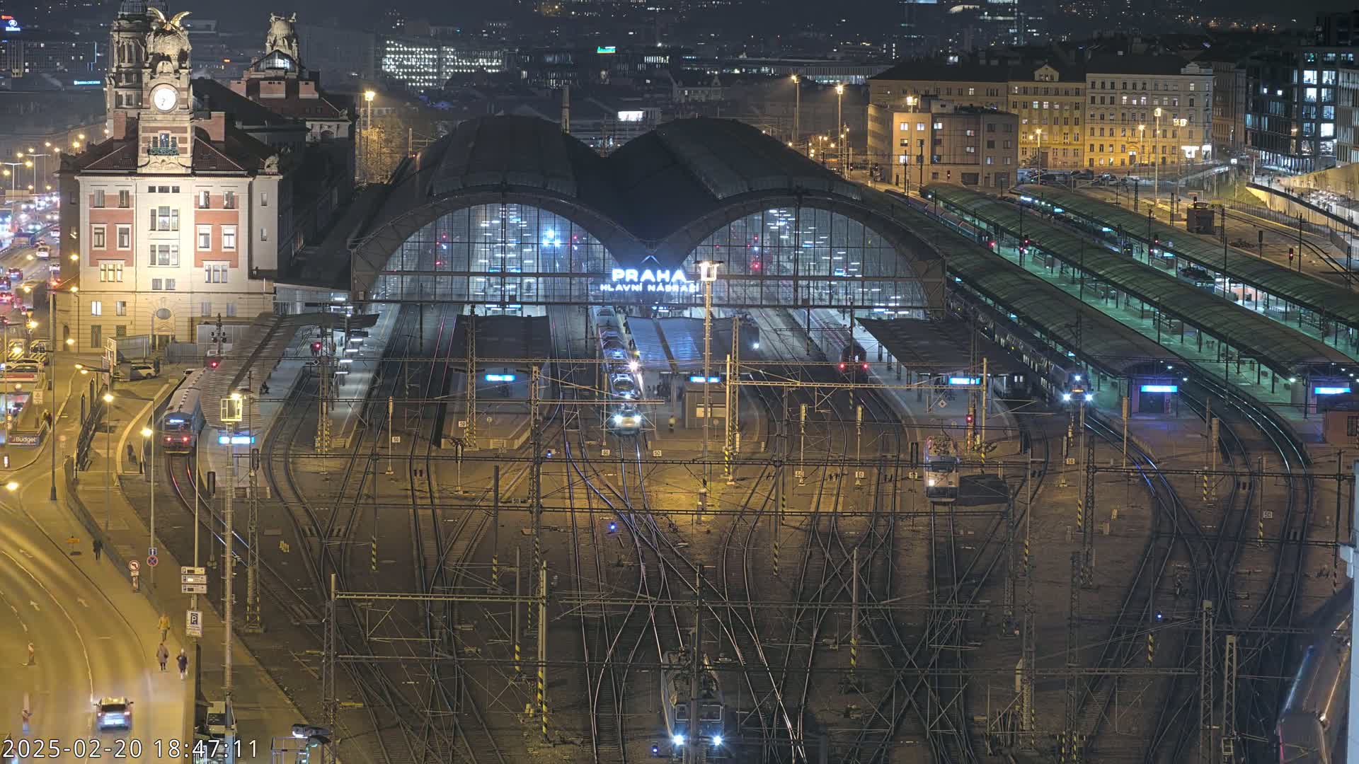 A nighttime aerial view of a large, illuminated train station with numerous tracks and trains, surrounded by city buildings and roads.