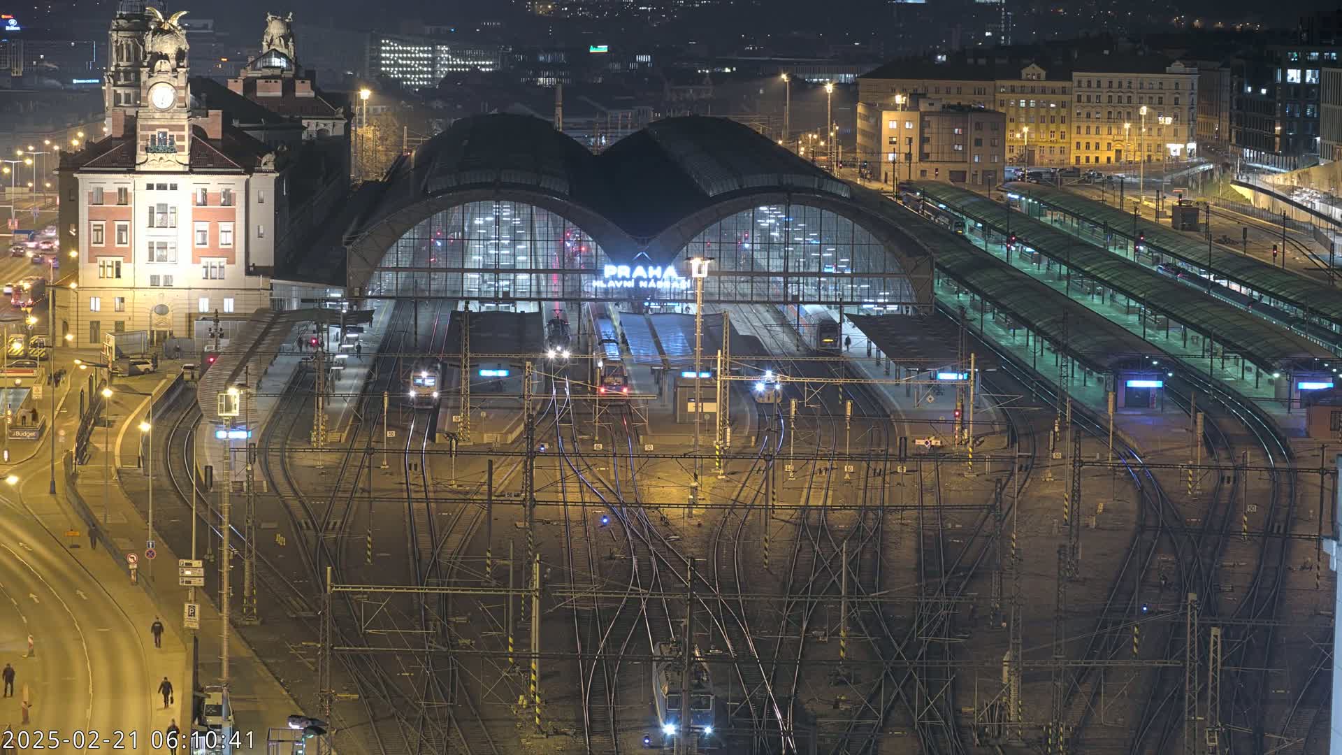 A nighttime aerial view of a large, illuminated train station with numerous tracks and platforms, surrounded by city buildings.