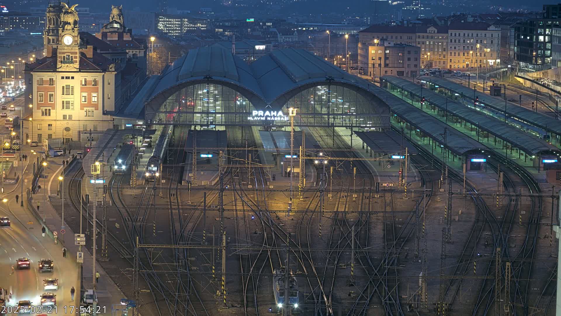 A large, illuminated train station at night, surrounded by a complex network of train tracks and city streets with cars driving on them.