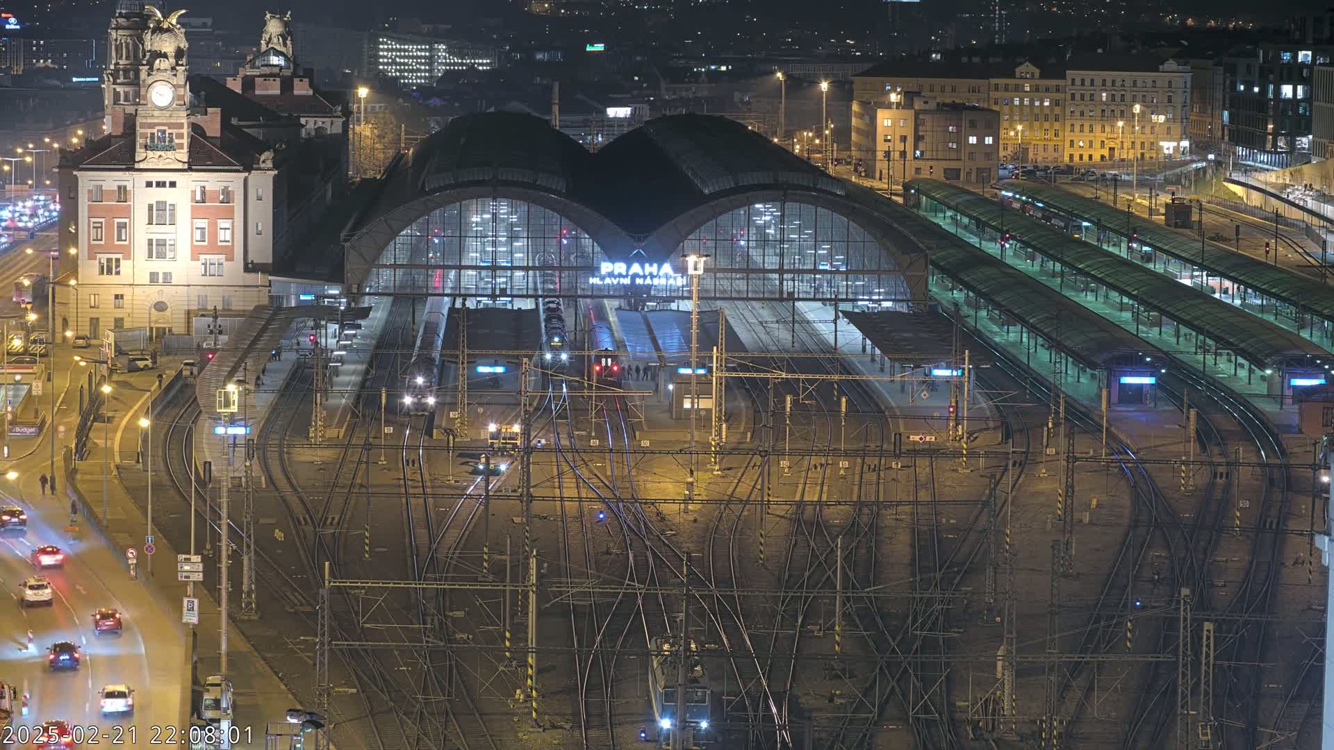 A nighttime, high-angle view of a busy train station with numerous tracks, platforms, and a large building, illuminated by artificial lights.