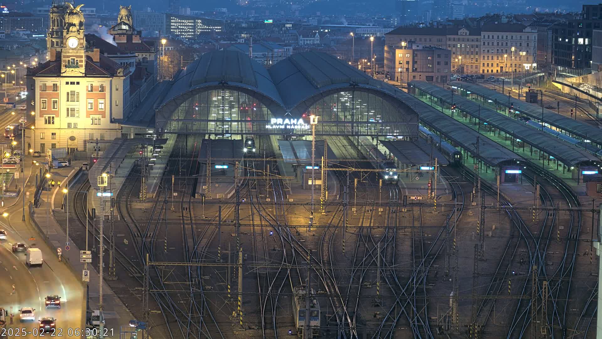 A nighttime aerial view of a large, busy train station with many tracks and platforms, surrounded by city buildings under a dark sky.
