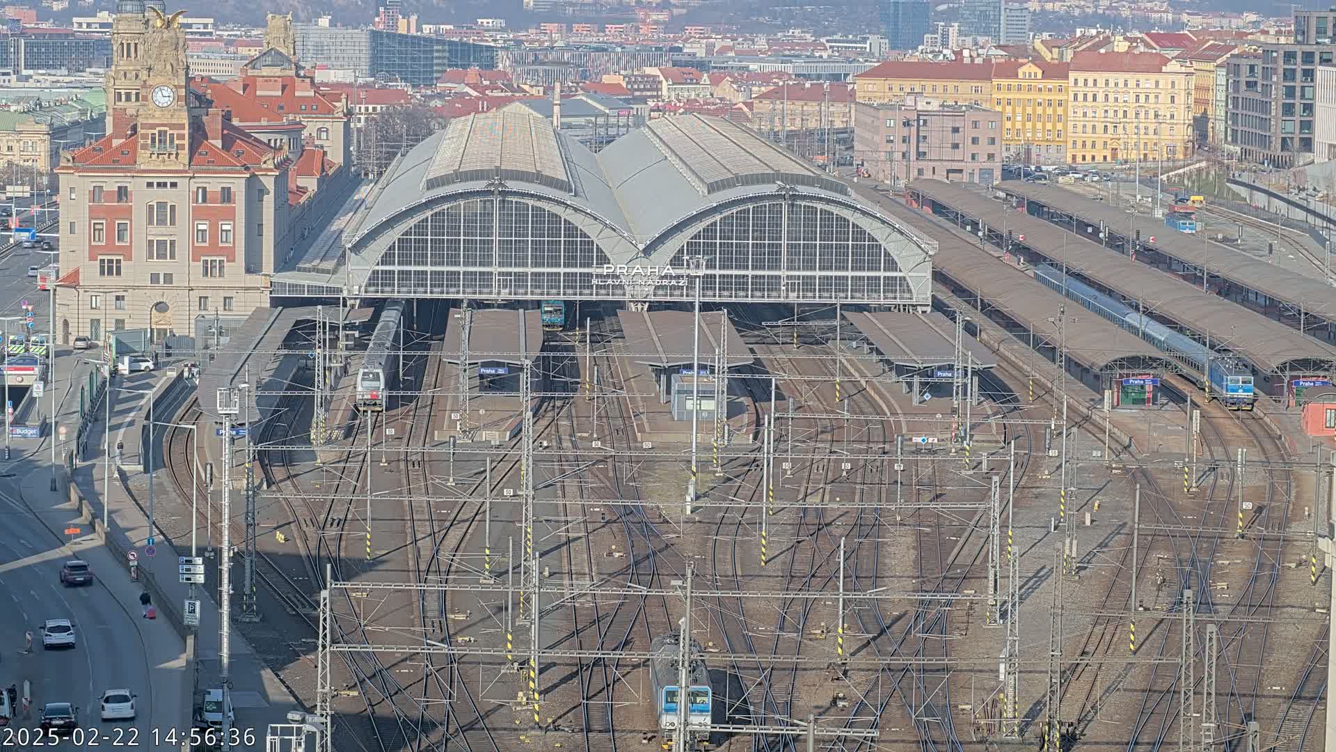 A large train station with numerous tracks and platforms, a substantial brick building, and several trains, all under a sunny sky.