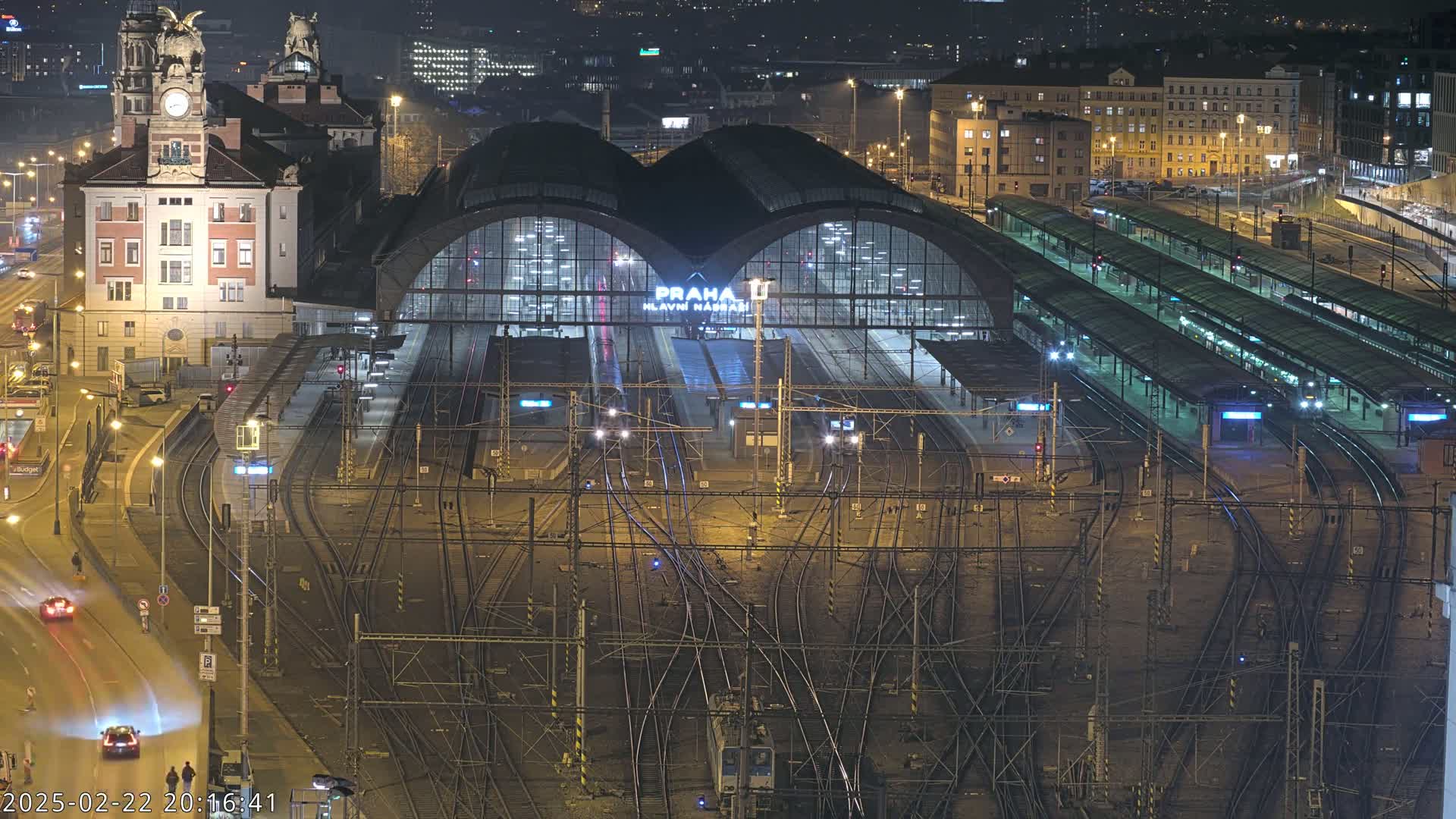 A large, illuminated train station at night, with numerous tracks and platforms extending from a central building, surrounded by city lights and a few cars driving on a nearby road.
