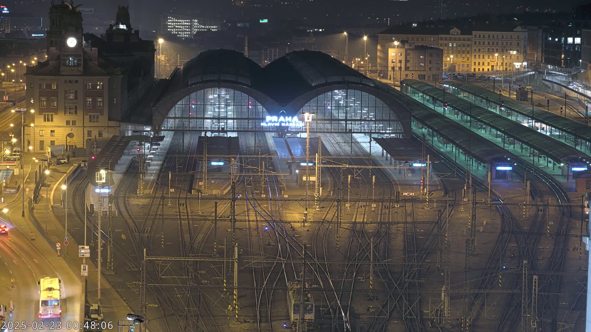 A nighttime aerial view of a large, illuminated train station with numerous tracks and platforms, surrounded by city buildings.