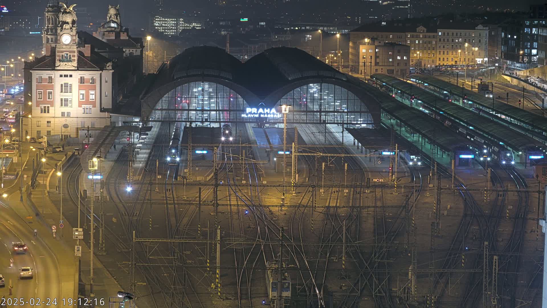 A nighttime aerial view of a large, illuminated train station with numerous tracks and platforms, surrounded by city buildings.