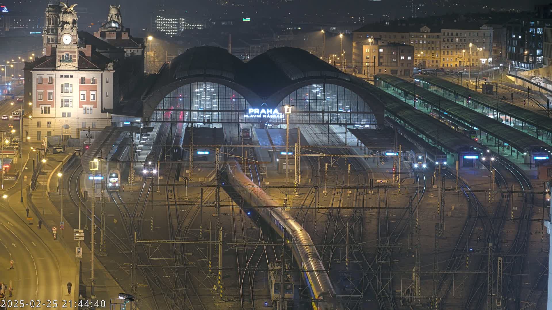 A nighttime aerial view of a large, illuminated train station with numerous tracks and trains, surrounded by city buildings.