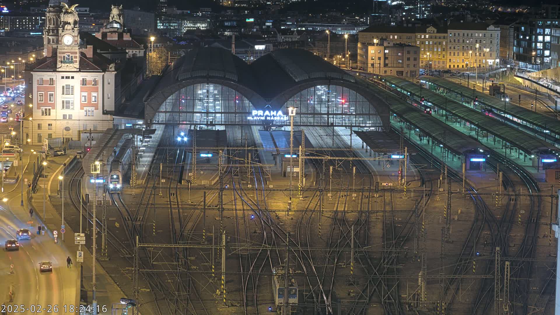 A nighttime aerial view of a large, illuminated train station with numerous tracks and platforms, surrounded by city buildings and a road with moving vehicles.
