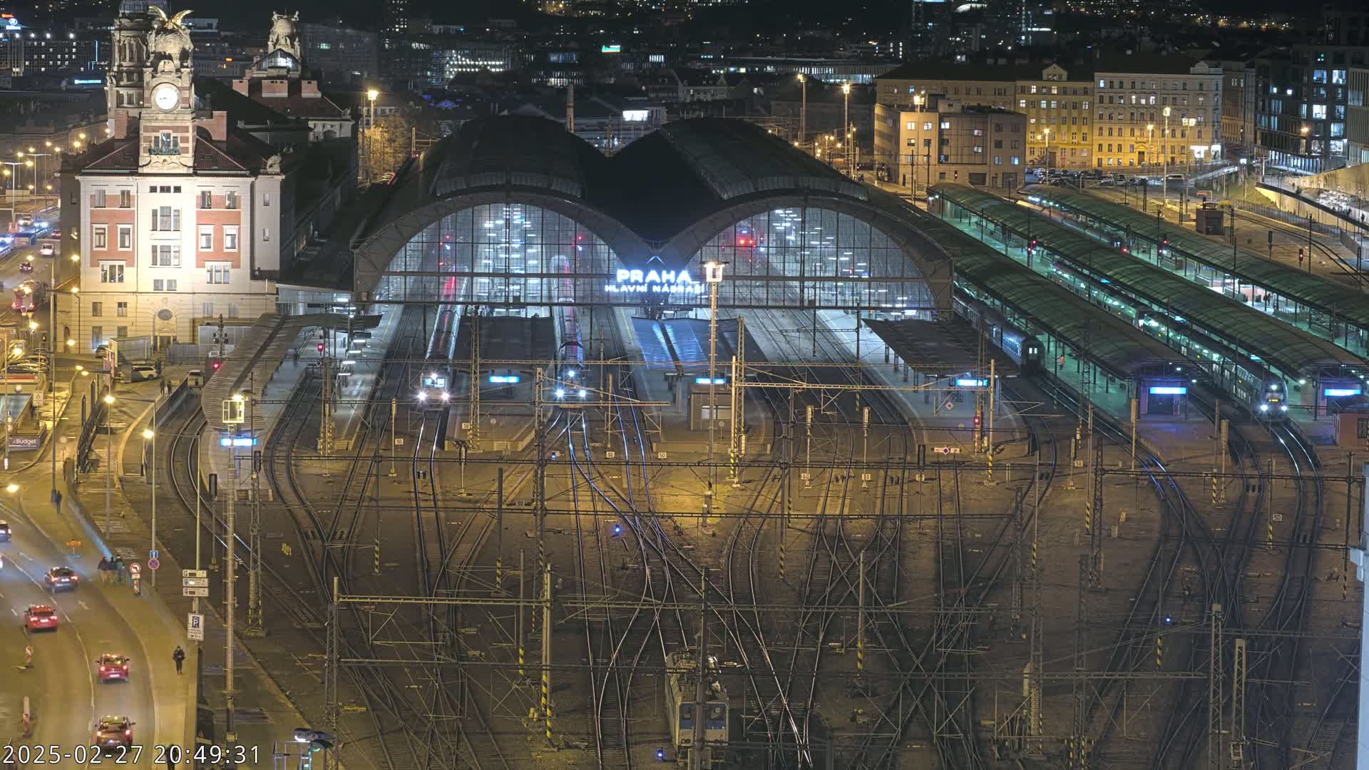 A nighttime aerial view of a large, illuminated train station with many tracks and platforms, surrounded by city buildings and roads with cars.