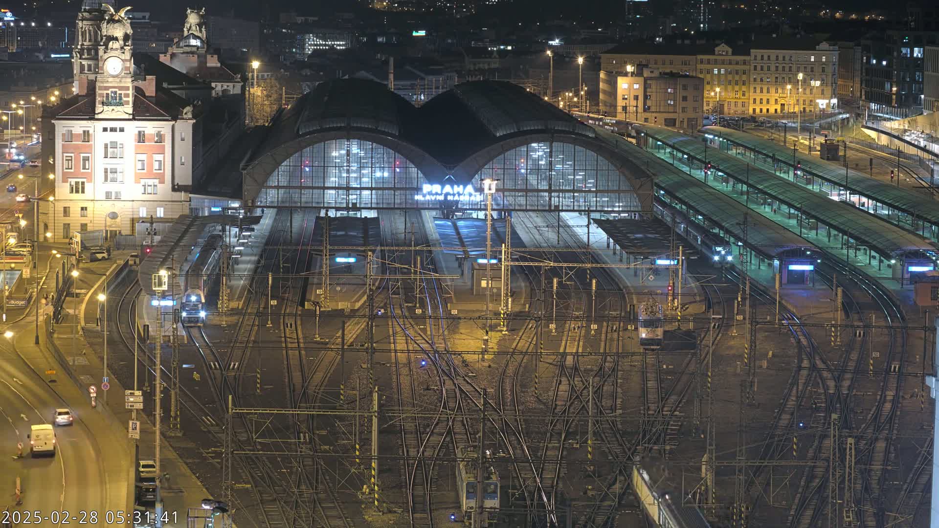 A nighttime aerial view of a large, illuminated train station with numerous tracks and trains, surrounded by city buildings.