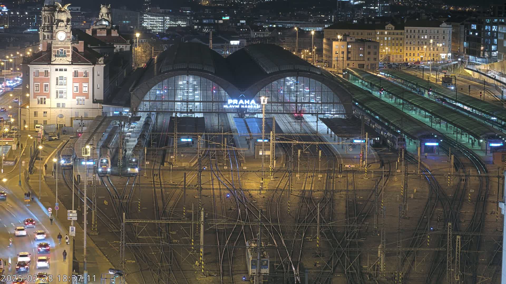 A large, illuminated train station at night, with numerous tracks and trains, is surrounded by city buildings.