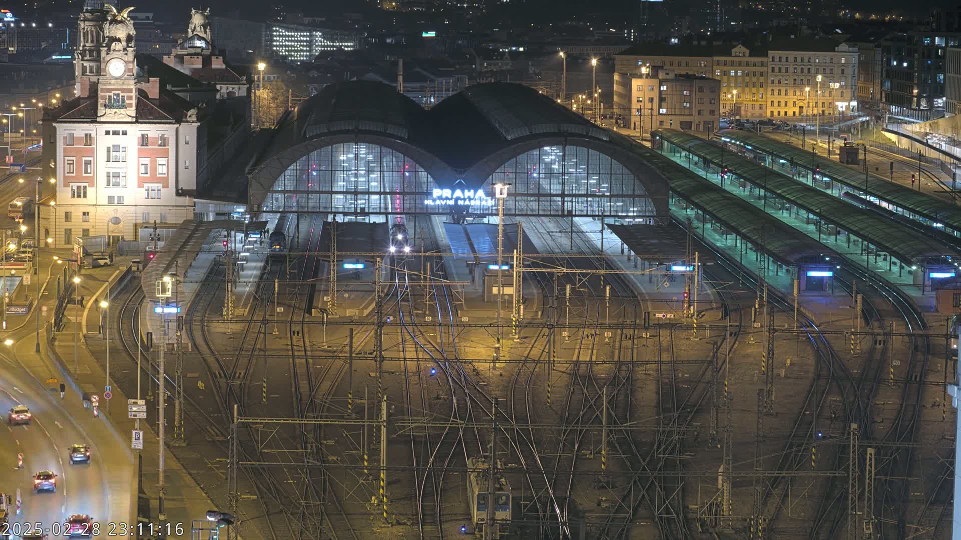 A nighttime aerial view of a large, illuminated train station with many tracks and platforms, surrounded by city buildings.