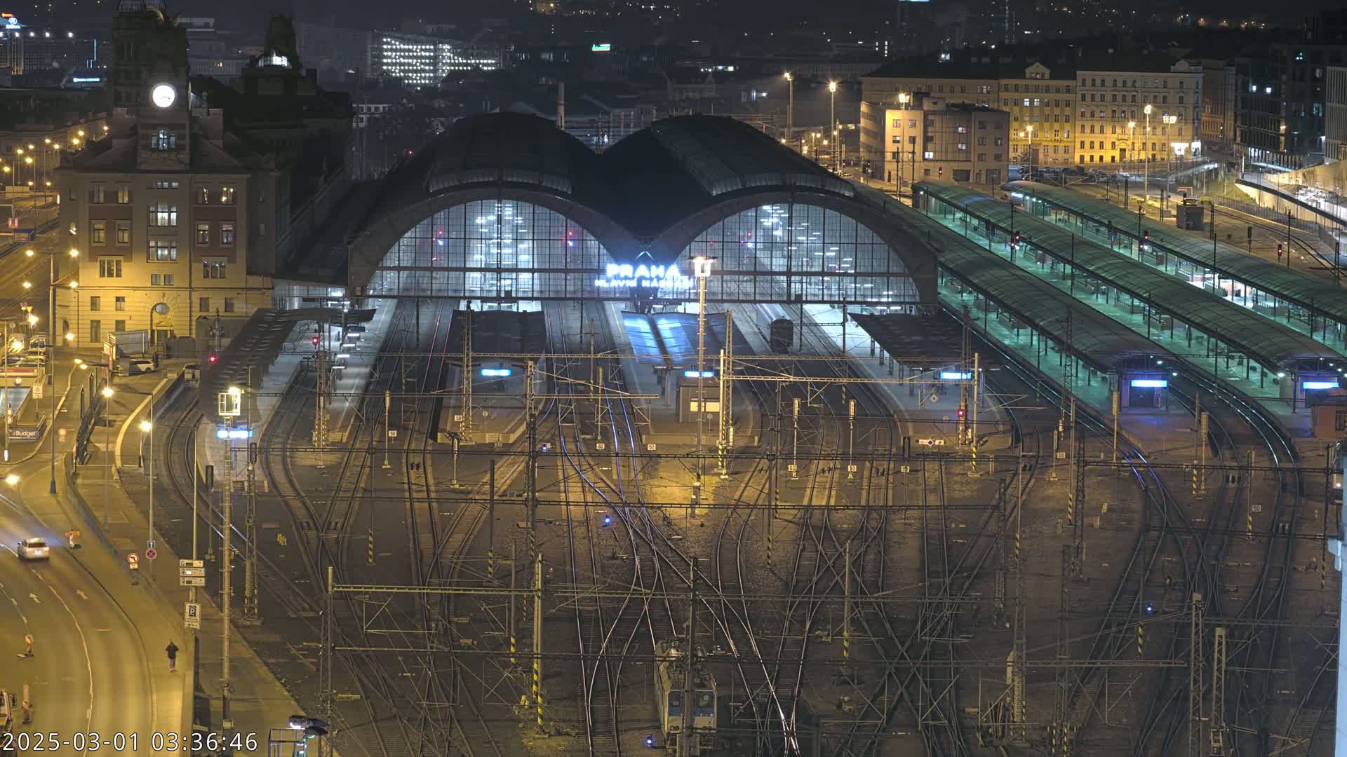 A nighttime aerial view of a large, illuminated train station with numerous tracks and platforms, surrounded by city buildings.
