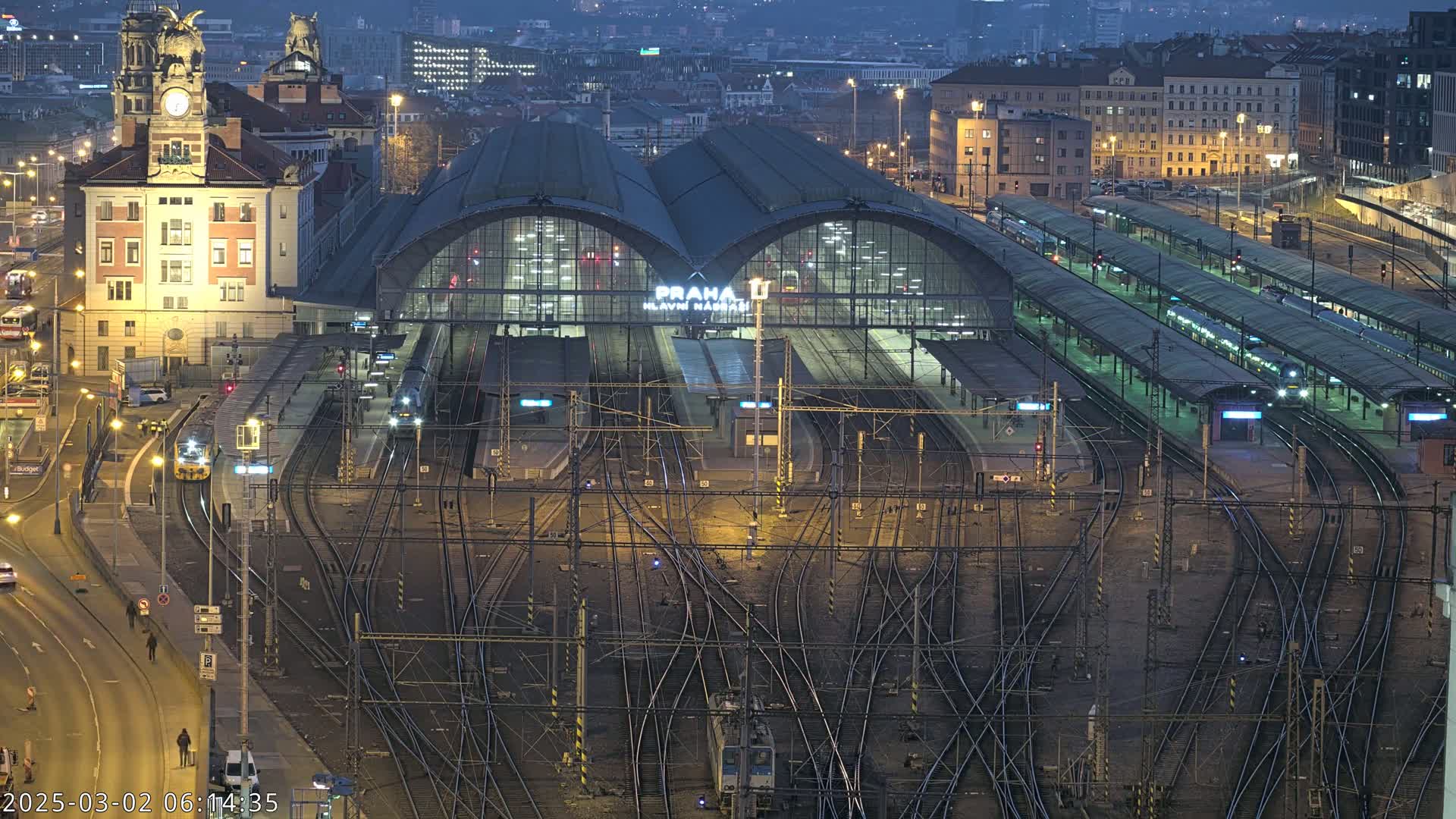 A large, illuminated train station at night with numerous tracks and trains, surrounded by city buildings under a dark sky.