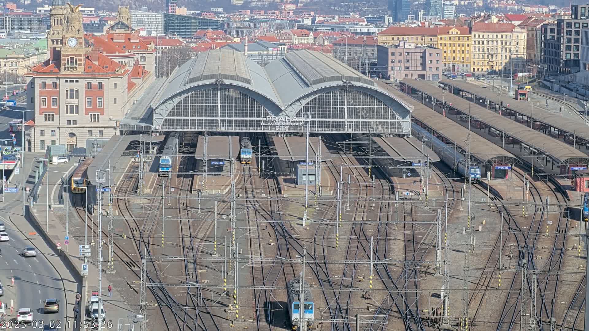 A large train station with many tracks and several trains, under a sunny sky, is surrounded by city buildings.