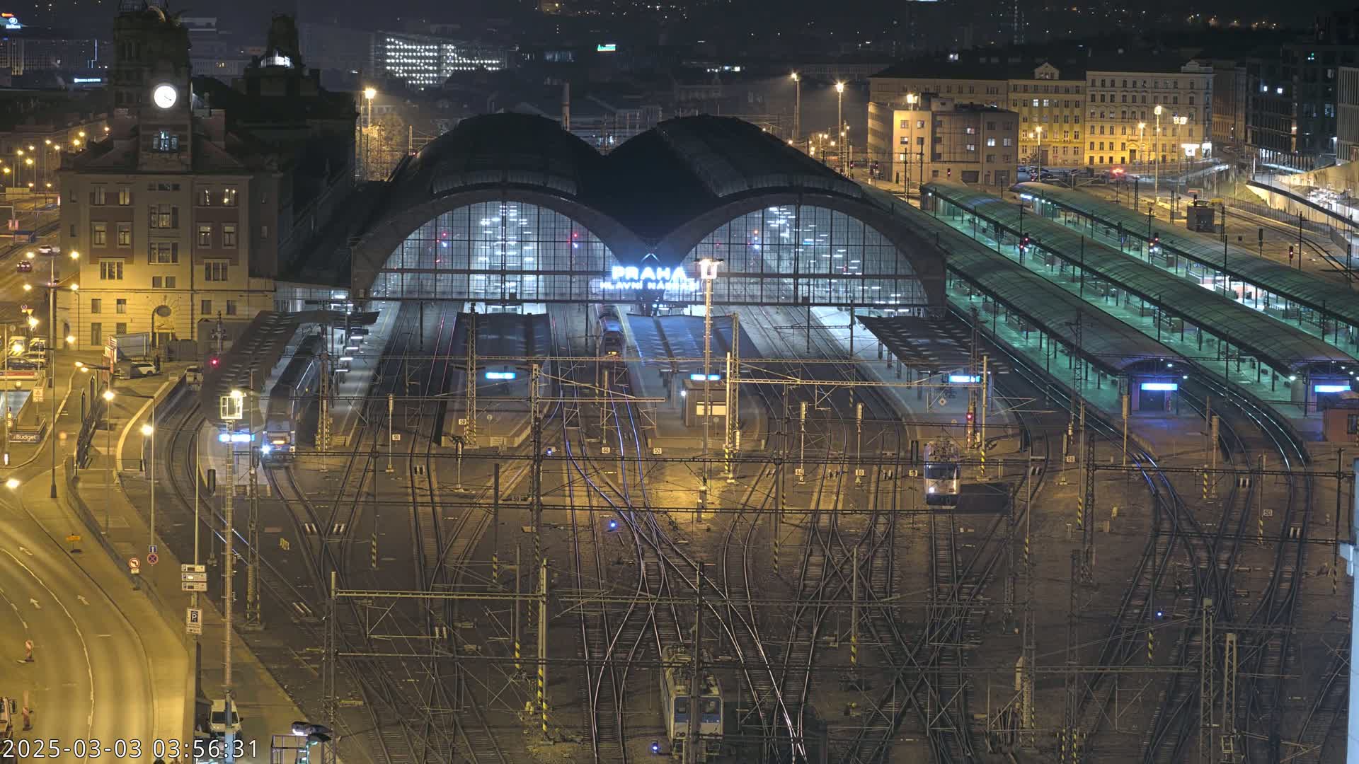 A nighttime aerial view of a large, illuminated train station with numerous tracks and trains, surrounded by city buildings.