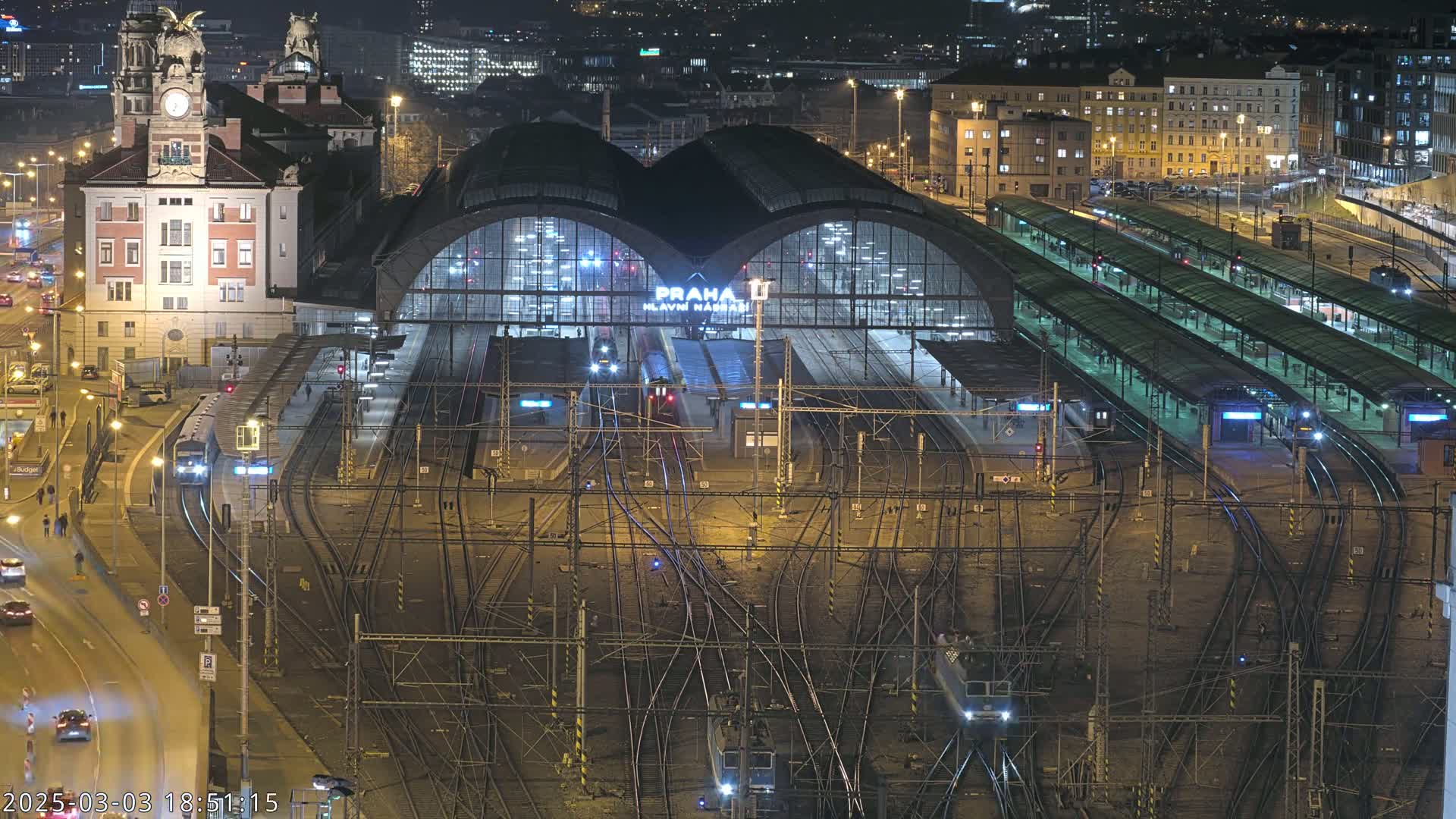 A nighttime aerial view of a large train station with numerous tracks, platforms, and trains, surrounded by city buildings under clear skies.