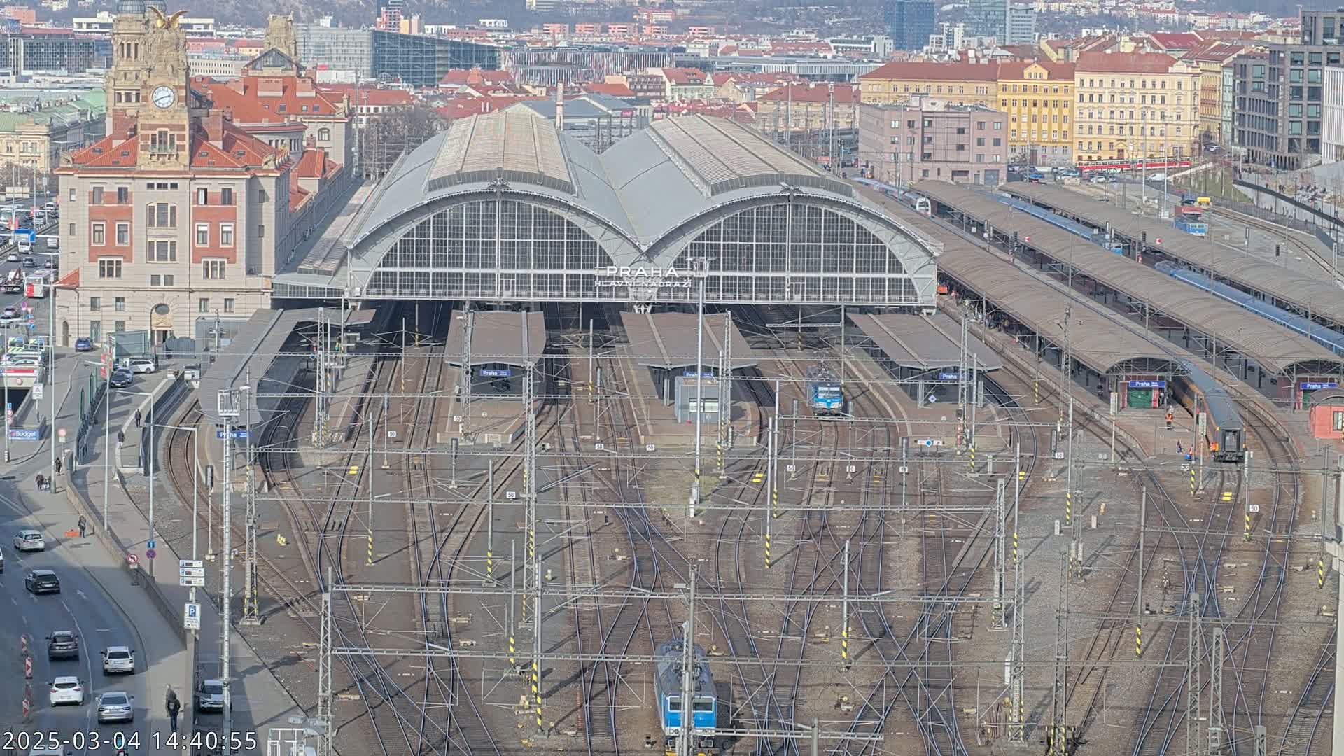 A large train station with numerous tracks and platforms, surrounded by city buildings under a sunny sky.