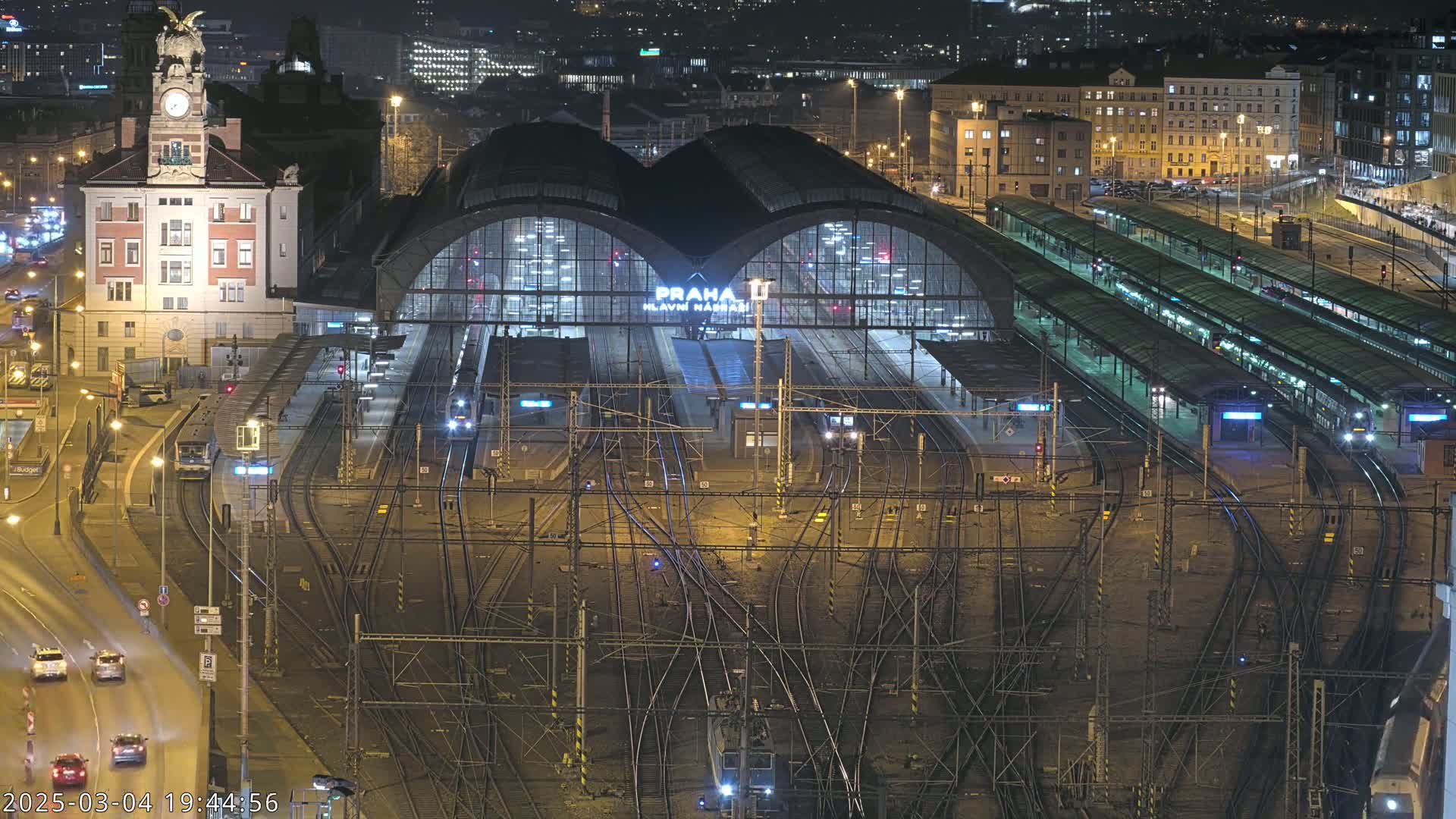 A nighttime aerial view of a large, illuminated train station with numerous tracks and trains, surrounded by city buildings.