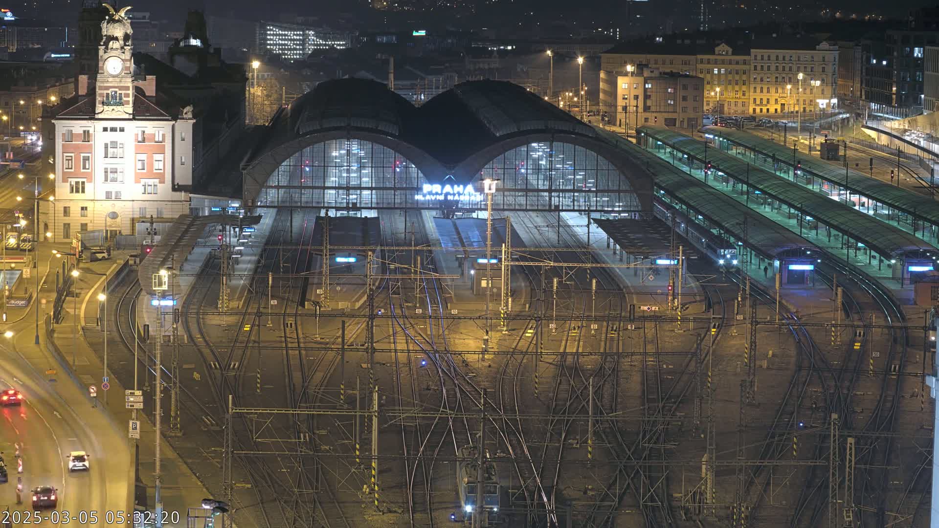 A nighttime aerial view of a large, illuminated train station with numerous tracks and platforms, surrounded by city buildings.