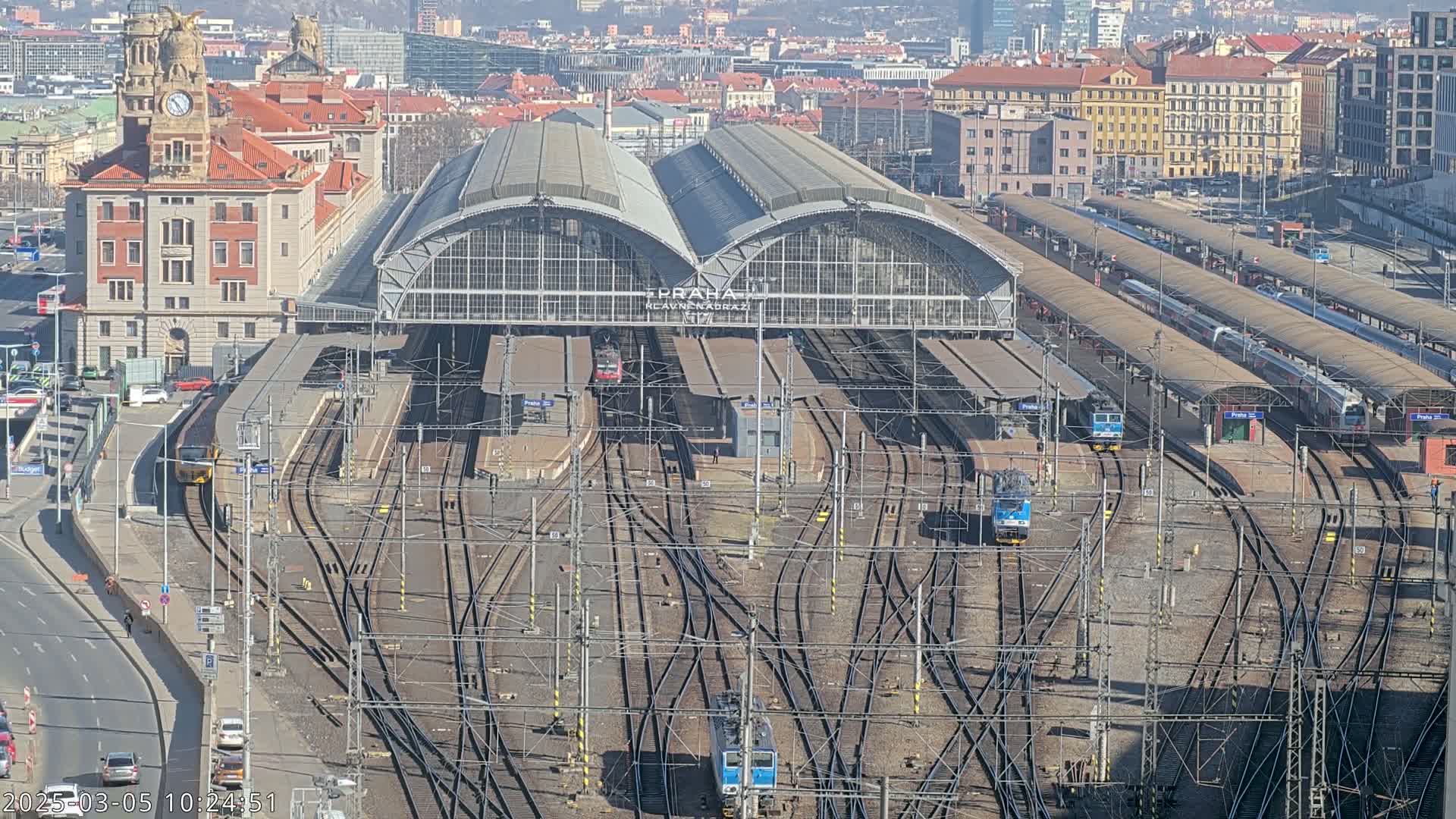 A large train station with numerous tracks and trains, under a sunny sky, is viewed from above.