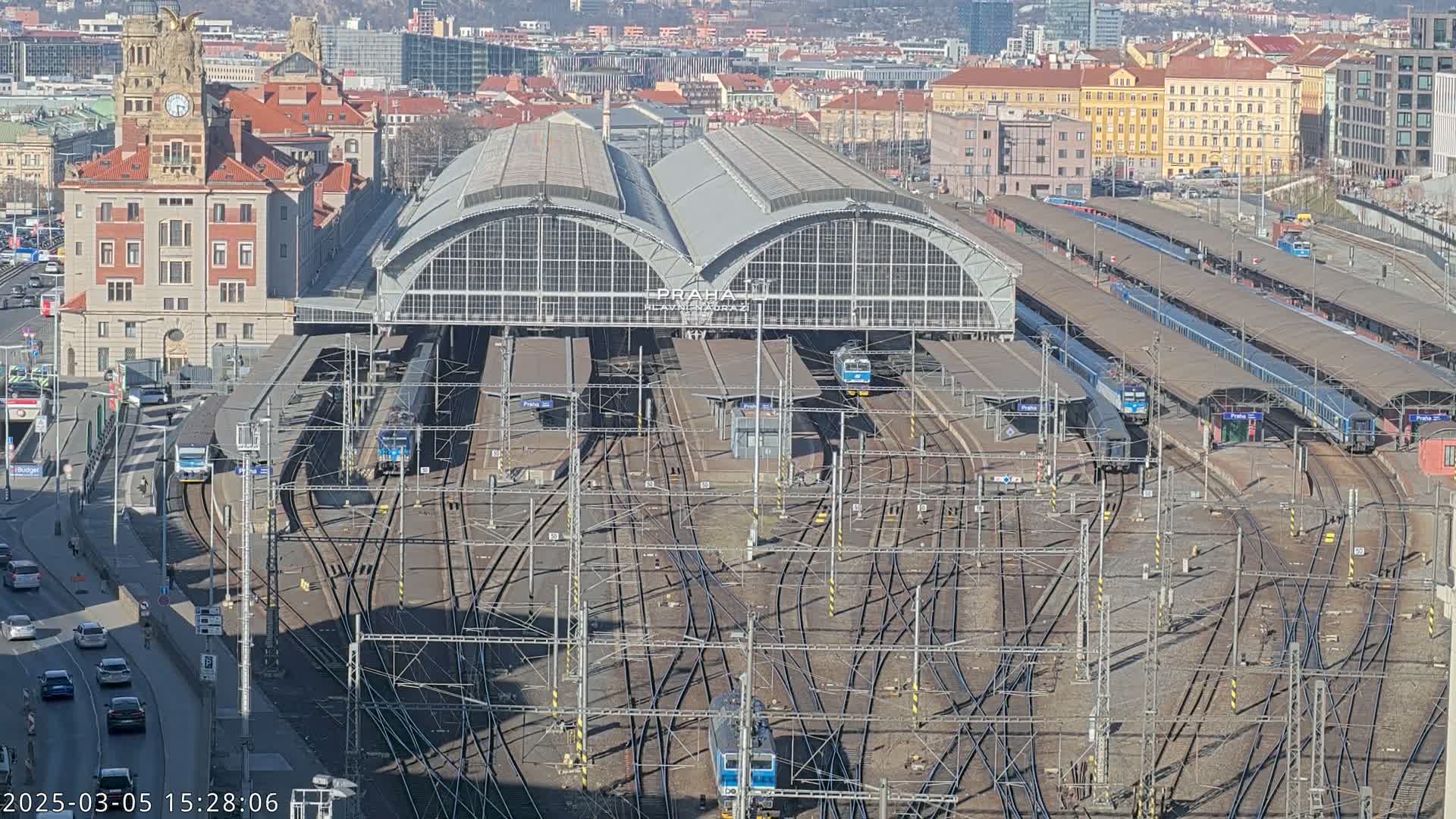 A large train station with numerous tracks and trains, under a sunny sky, is surrounded by a city.