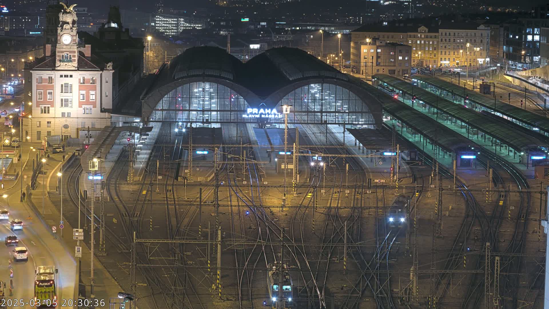 A nighttime aerial view of a large, illuminated train station with numerous tracks and platforms, surrounded by city buildings.