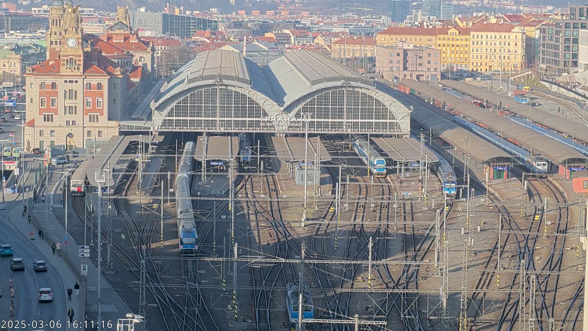 A large train station with numerous trains on tracks, surrounded by city buildings under a sunny sky.
