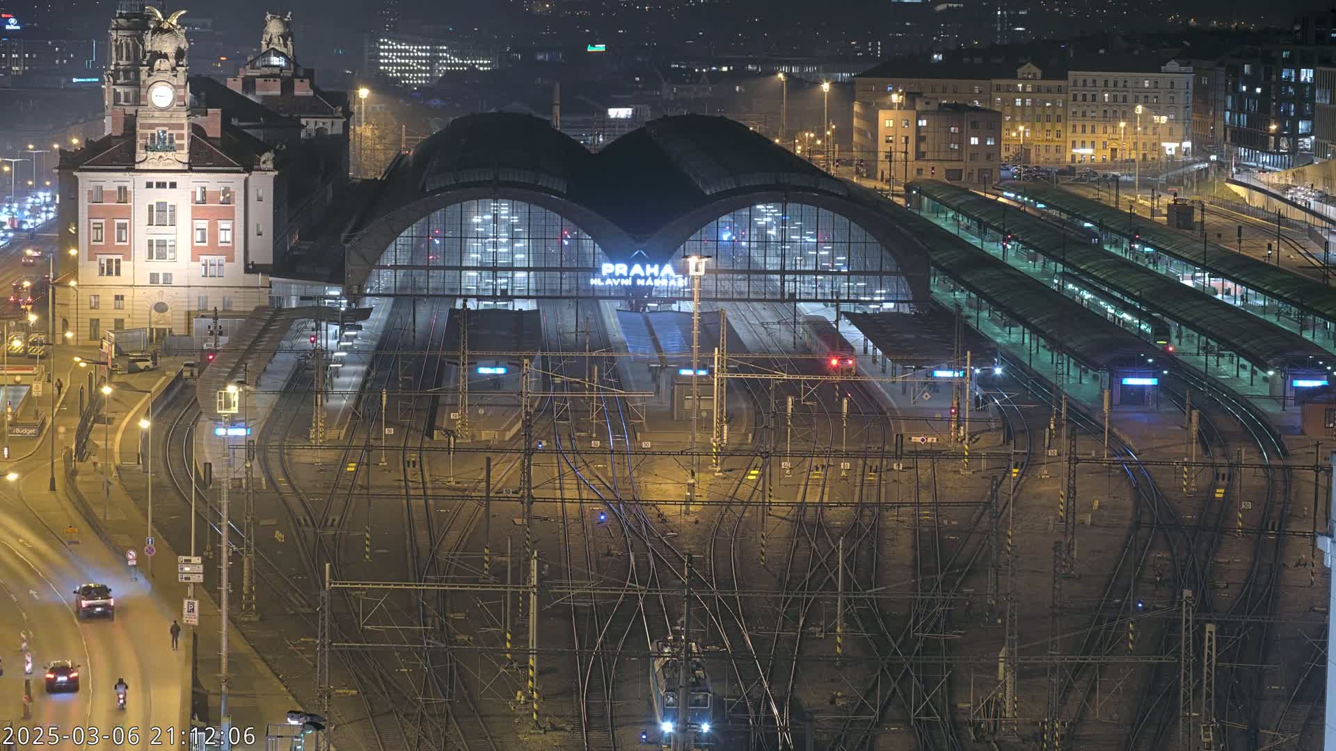 A nighttime aerial view of a large, illuminated train station with numerous tracks and platforms, surrounded by city buildings under clear skies.