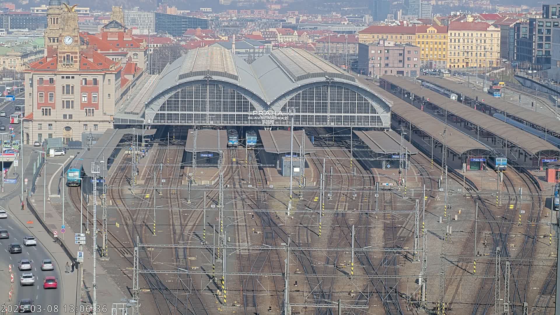A large train station with numerous tracks and platforms, under a clear sunny sky, is surrounded by a city.