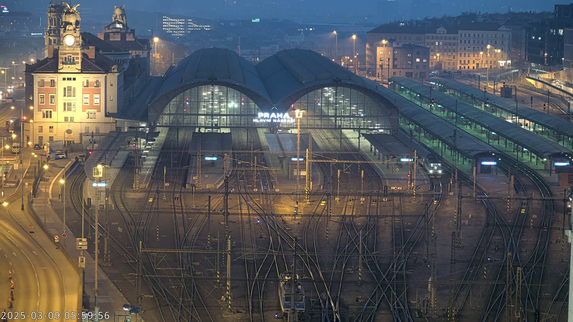 A nighttime, high-angle view of a large, illuminated train station with numerous tracks and platforms, surrounded by city buildings.