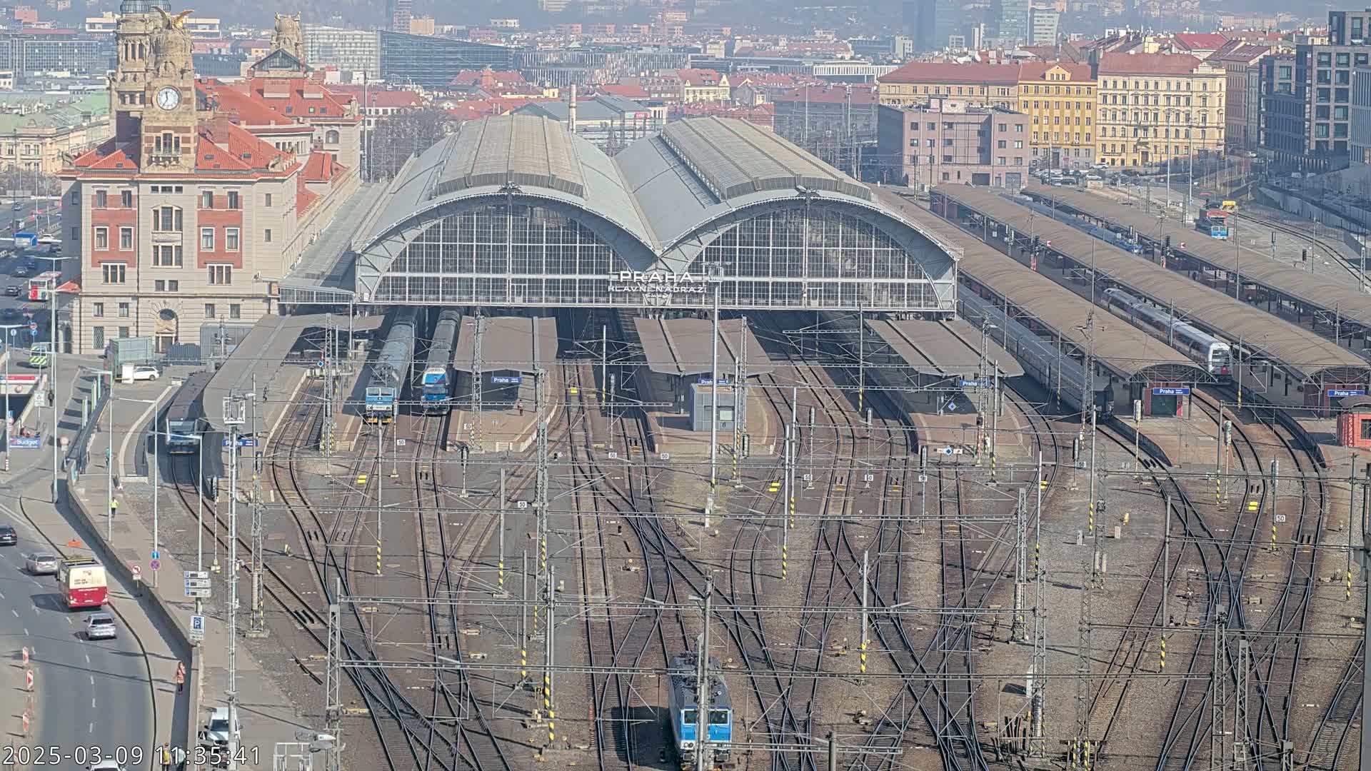 A large train station with numerous tracks and several trains, viewed from above on a sunny day.