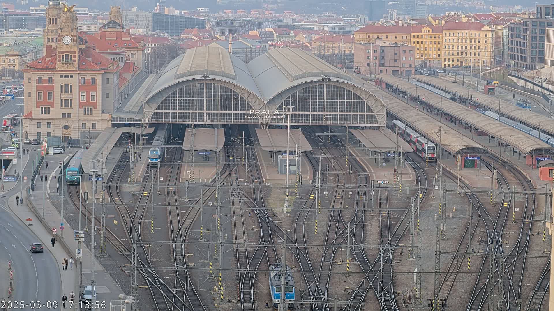 A large train station with numerous tracks and platforms, surrounded by city buildings under an overcast sky.
