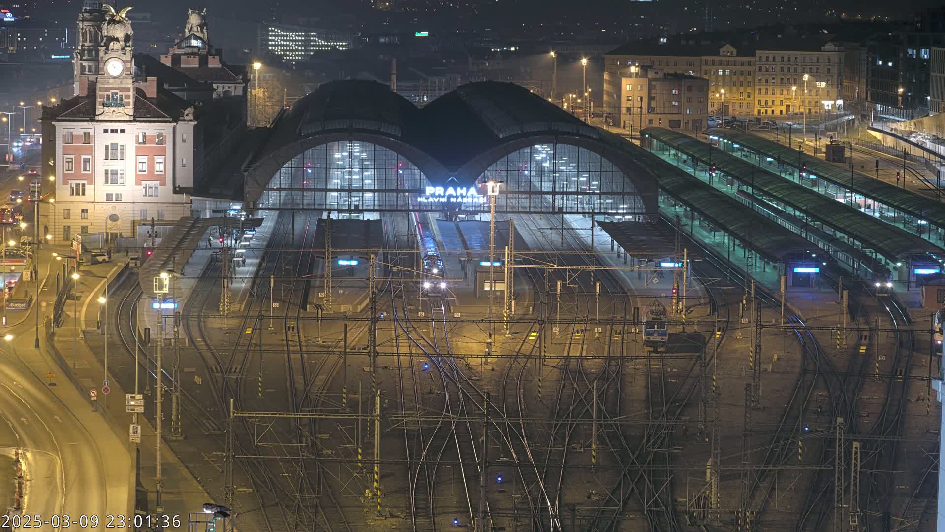 A brightly lit train station at night, with many train tracks and platforms extending from a large central building, surrounded by city buildings under a dark sky.