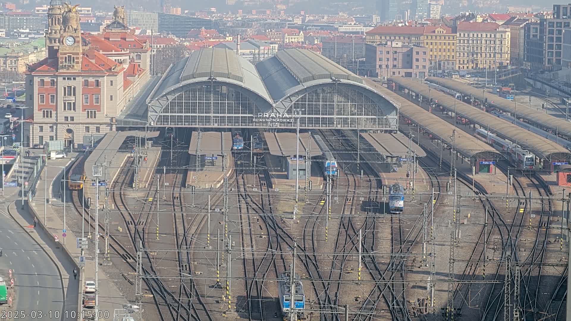 A large train station with numerous tracks and trains, situated in a city under a hazy, sunny sky.