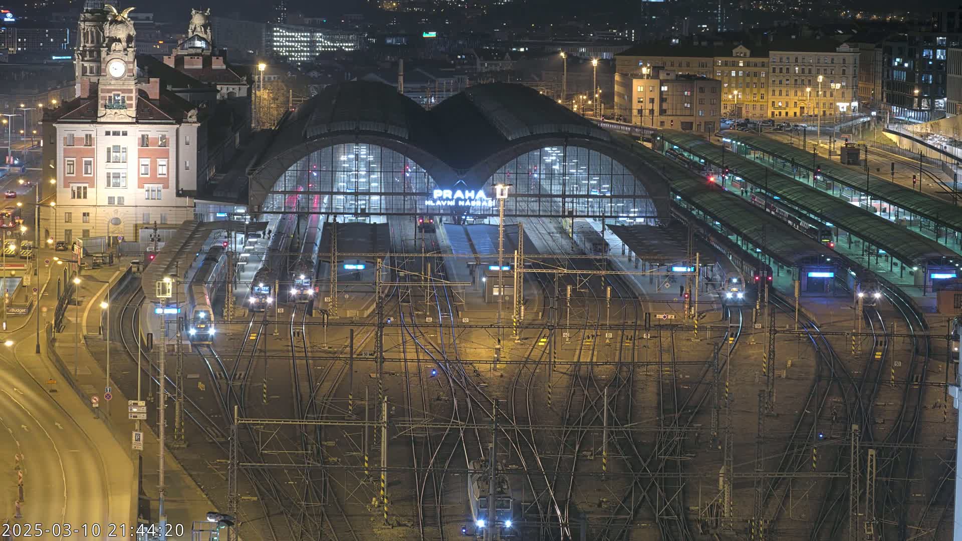 A large, illuminated train station at night, with numerous trains on its many tracks, surrounded by city buildings.