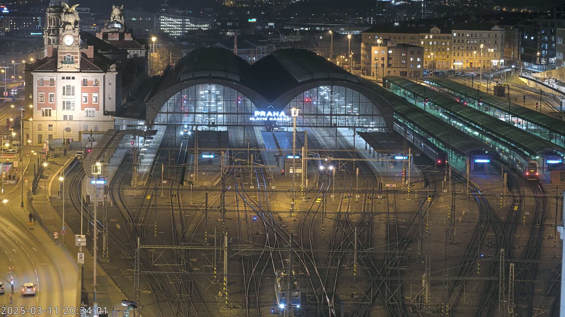 A large, illuminated train station at night, with numerous train tracks and platforms, surrounded by city buildings.