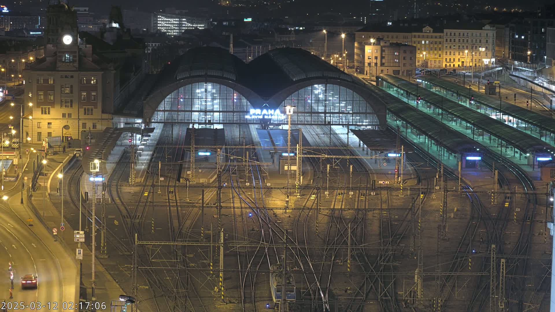 A brightly lit train station at night, with a complex network of train tracks surrounding it and a city skyline visible in the background.