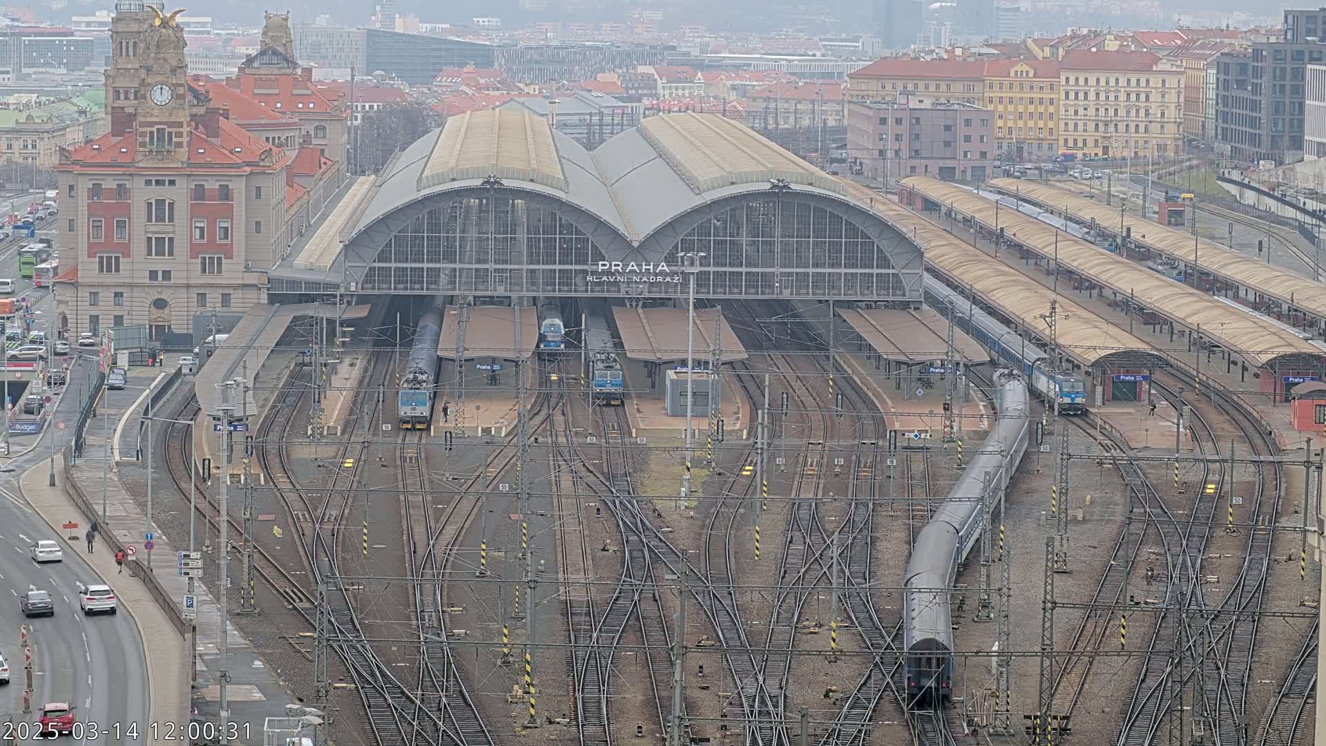 A large train station with many tracks and several trains, situated in a city on an overcast day.