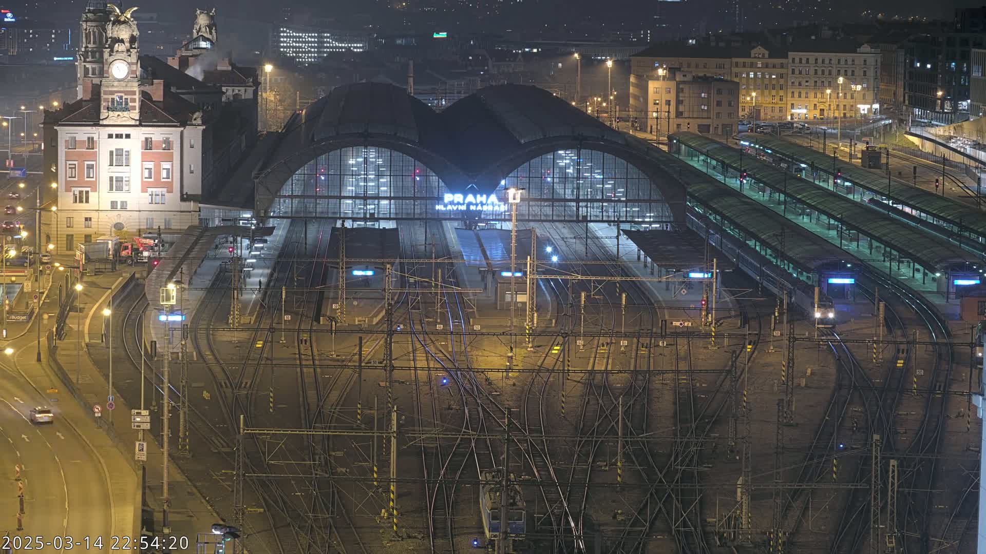 A nighttime view of a large, illuminated train station with numerous tracks and trains, surrounded by city buildings.