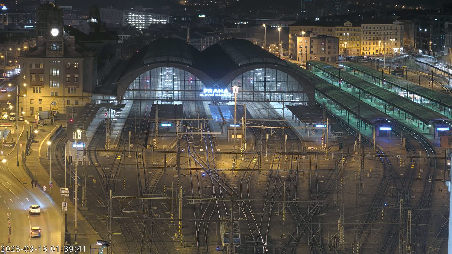 A nighttime aerial view of a large, illuminated train station with numerous tracks and platforms, surrounded by city buildings.