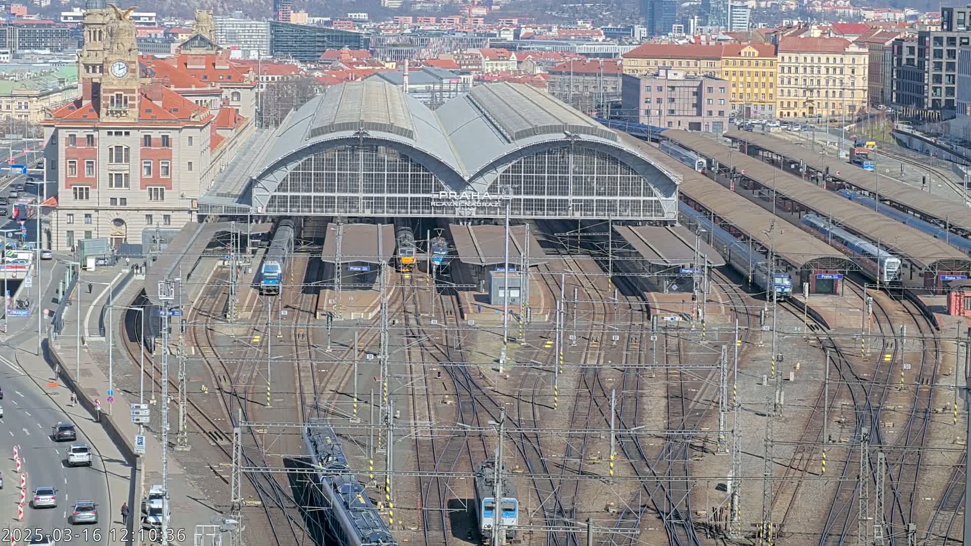 A large train station with numerous trains and tracks, surrounded by city buildings under a sunny sky.